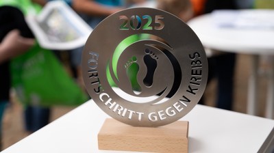 Rundes Metallobjekt mit ausgeschnittenen Fußabdrücken und der Aufschrift '2025 FORTSCHRITT GEGEN KREBS' auf einem Holzsockel.