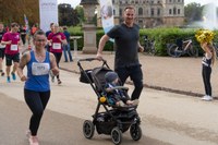 Läuferin mit Startnummer 1073 und Mann, der Kinderwagen mit Kind schiebt, laufen auf einem Weg in einem Park, im Hintergrund weitere Läufer und eine Person mit goldfarbenen Pompons