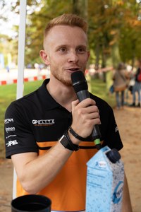 Junger Mann mit kurzem roten Haar spricht in ein Mikrofon, trägt ein schwarz-orangefarbenes Polohemd mit mehreren Logos, im Hintergrund unscharfe Personen und Bäume.