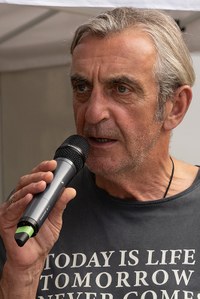 Älterer Mann mit grauen Haaren spricht in ein Mikrofon und trägt ein T-Shirt mit der Aufschrift 'TODAY IS LIFE TOMORROW IS'