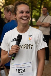 Lächelnde Frau mit Mikrofon trägt ein weißes T-Shirt mit der Aufschrift "Germany" und einer Startnummer mit dem Namen "Saskia" bei einer Veranstaltung im Freien