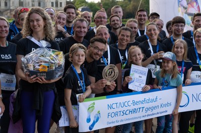 Gruppe von Menschen mit Medaillen und Urkunden bei einer Benefizveranstaltung gegen Krebs, einige halten einen Korb und eine Trophäe, Banner mit Text zur Unterstützung des NCT/UCC-Benefizlaufs