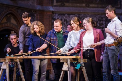 (c) NCT/UCC (André Wirsig) Gruppe von sieben jungen Menschen steht um ein langes Holzbrett mit Nägeln auf zwei Böcken und hält Bögen, einige blicken konzentriert auf das Brett