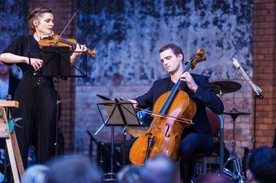 (c) NCT/UCC (André Wirsig) Eine Frau spielt Geige und ein Mann spielt Cello vor einem gemauerten Hintergrund bei einem Konzert.
