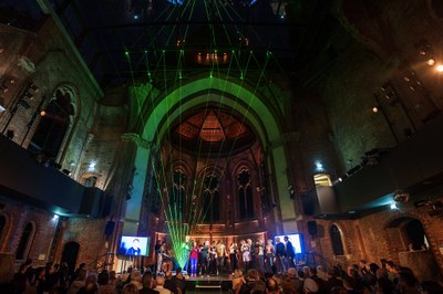 (c) NCT/UCC (André Wirsig) Menschen stehen auf einer Bühne in einer großen, historischen Halle mit Rundbögen und Ziegelwänden, grüne Laserstrahlen sind sichtbar, Publikum sitzt davor
