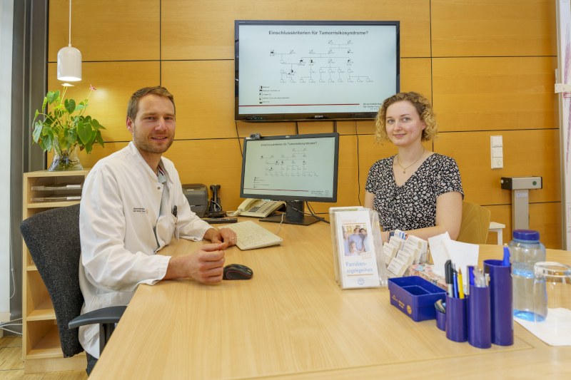 Two people sitting at a table in an office with two screens displaying genetic diagrams in the background
